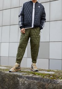 Vadderad svart bomberjacka med vita diagonala ränder, olivgröna vadderade joggers och beige sneakers med kamouflage-mönster.