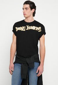 Μαύρο βαμβακερό T-shirt με κοντά, γυρισμένα μανίκια και ένα έντονο χρυσό τυπωμένο λογότυπο στη μέση του στήθους. Φορεμένο με μπλε τζιν και μαύρο φούτερ δεμένο στη μέση.