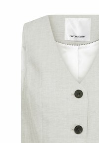 Vaaleanharmaa liivi, jossa on V-pääntie, herringbone-kudonta ja mustat napit. Sisällä etiketti lukee "co'couture."