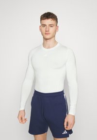 Wit sportshirt met lange mouwen en marineblauwe shorts. Het shirt heeft een gladde textuur en een ronde hals, met een klein logo op de borst.