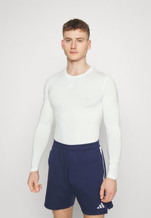 Weißes langärmliges Sportoberteil und marineblaue Shorts. Das Oberteil hat eine glatte Textur und einen Rundhalsausschnitt, mit einem kleinen Logo auf der Brust.