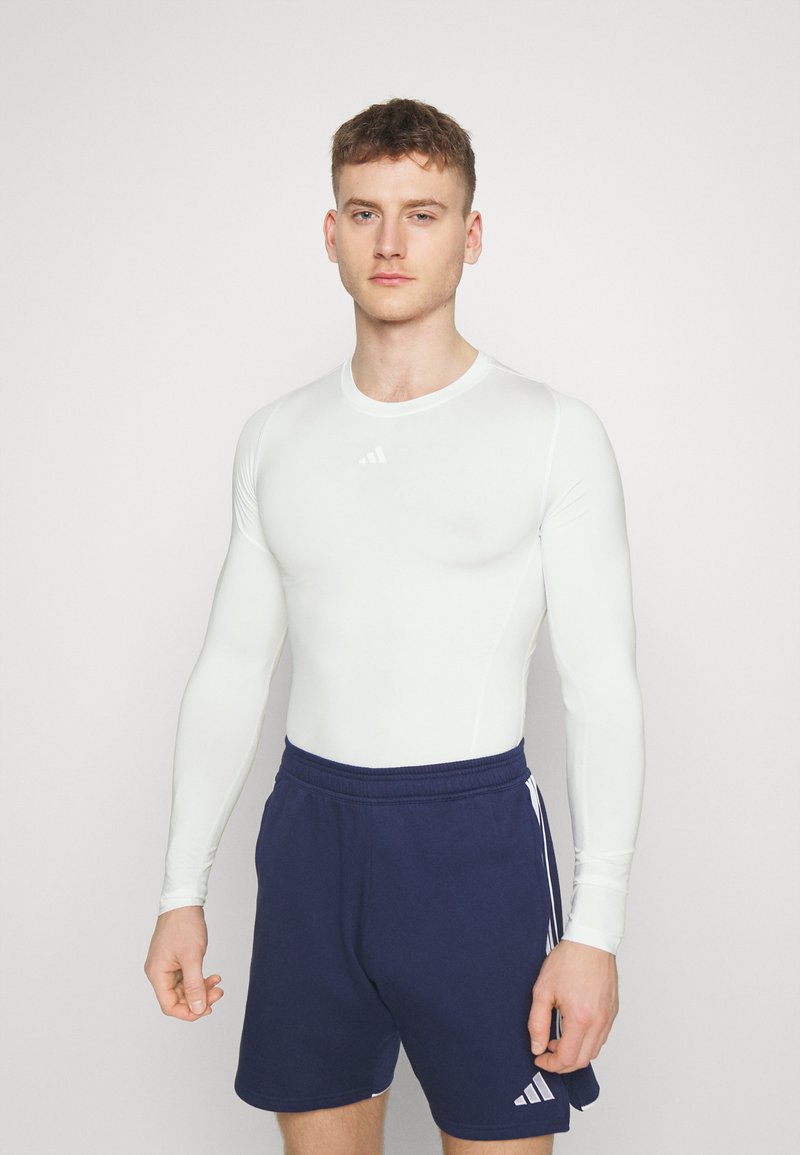 Wit sportshirt met lange mouwen en marineblauwe shorts. Het shirt heeft een gladde textuur en een ronde hals, met een klein logo op de borst.