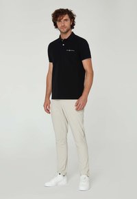 Polo negro de manga corta, con cuello y detalles en azul claro. Combinado con pantalones beige y zapatillas blancas en una pose de pie.