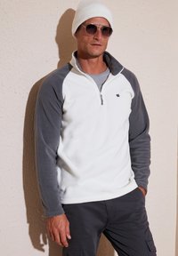 Hvid og grå fleece-pullover med halv-zip krave, raglanærmer og lille logo. Kombinationen med en hvid hue og solbriller.