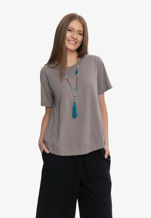 Uśmiechnięta kobieta z długimi prostymi włosami, ubrana w szary krótki top, czarne szerokie spodnie i niebieski naszyjnik z koralików z frędzlami.