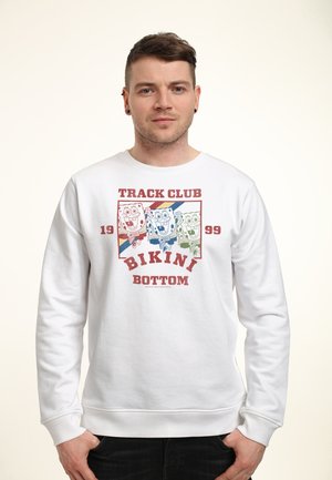 Man met wit sweatshirt met cartoon SpongeBob-personages en tekst "Track Club Bikini Bottom 1999" aan de voorkant, staand met handen in de zakken.