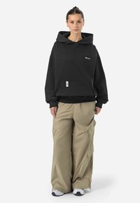 Svart huvtröja med framficka, matchad med lösa beige cargobyxor. Modellen bär vita sneakers; minimalistisk design med logodetalj.