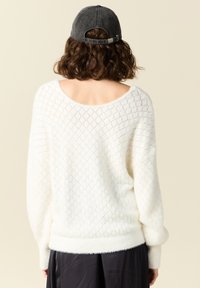 Maglione bianco e peloso lavorato a maglia con un motivo a rombi, scollatura ampia e polsini a coste. Indossato con un berretto grigio, dotato di una fascia sul retro.
