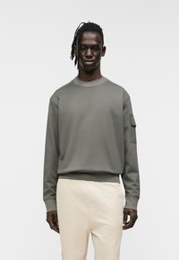 Φούτερ σε ελιά πράσινο με στρογγυλή λαιμόκοψη, ριμπ μανσέτες και τσέπη-μπατζάκι στο αριστερό μανίκι. Απαλή ύφανση και άνετη εφαρμογή.