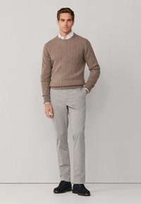 Brązowy sweter z dzianiny z wzorem w warkocze, szare spodnie i granatowe buty. Model stoi z rękami w kieszeniach na neutralnym tle.