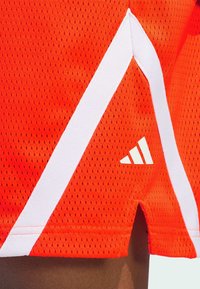 Červené síťované sportovní šortky s bílými akcentními pruhy a logem Adidas. Materiál je lehký s texturovaným povrchem.