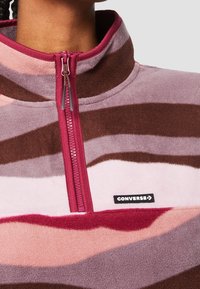 Primer plano de una persona que lleva un pulóver de forro polar a rayas en rosa, marrón y rojo, con una cremallera en color burdeos y un parche con el logo de Converse en el pecho.