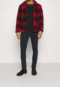 Röd och svart rutiga fleecejacka med två knappar, bärs över en svart skjorta, ihop med mörka byxor och svarta mocka ankelskort.