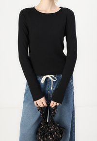 Schwarzes langärmliges Rippoberteil mit Rundhalsausschnitt, kombiniert mit lockeren blauen Jeans und einer gerüschten Tasche mit Blumenmotiv.