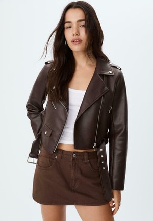 Junge Frau mit langen dunklen Haaren, die eine dunkelbraune Lederjacke, ein weißes Crop-Top und einen braunen Minirock vor einem einfarbigen hellen Hintergrund trägt.