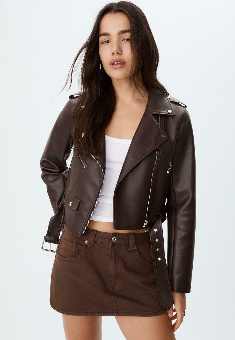 Giovane donna con lunghi capelli scuri, indossa una giacca di pelle marrone scuro, un crop top bianco e una mini gonna marrone, su uno sfondo chiaro e semplice.