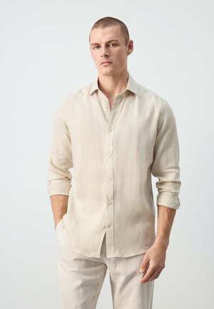 Homme aux cheveux courts portant une chemise beige à manches longues boutonnée et un pantalon clair assorti sur un fond uni.
