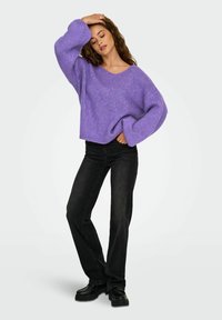 Donna in piedi con una mano sulla testa, che indossa un maglione oversized viola, jeans neri e scarpe nere su uno sfondo chiaro.