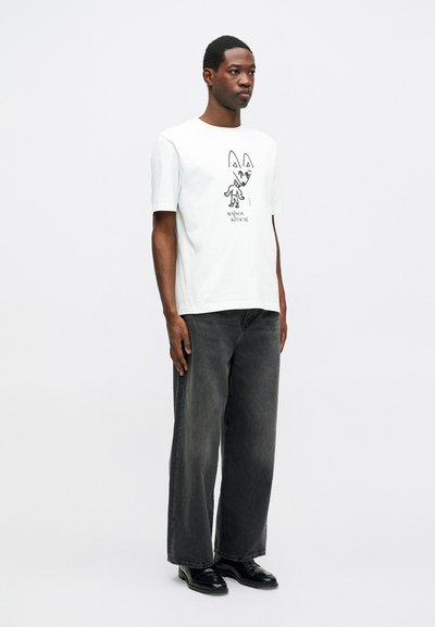 Muž v bílém grafickém tričku s designem lišky, volných tmavě šedých džínách a černých botách, stojící na jednobarevném bílém pozadí.