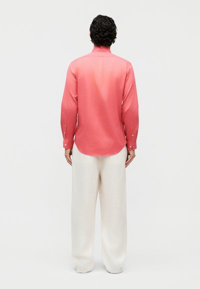 Άνδρας που στέκεται με την πλάτη στην κάμερα, φορώντας μακρυμάνικο κοραλί πουκάμισο και χαλαρό λευκό παντελόνι μπροστά από ένα απλό φόντο.