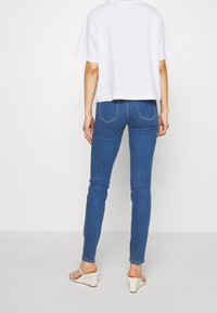 Osoba stojící zády, oblečená v bílém tričku s krátkým rukávem, modrých úzkých džínách a bílých sandálech na klínu na jednobarevném pozadí.