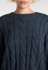 Navy blue cable-knit sweater featuring a textured design with twisting patterns and a wide neckline. Made from a soft knit material.