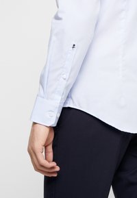 Blauw en witte gestreepte overhemd met knoopmanchetten, voorzien van een kleine zwarte rozenborduursel op de mouw. Glad stoftextuur.