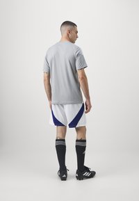 Camiseta deportiva gris, pantalones cortos blancos con acentos azules, calcetas negras hasta la rodilla con rayas blancas y botas de fútbol negras.