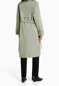Długi, jasnziezielony płaszcz typu trench wykonany z gładkiej tkaniny, z paskiem w talii i mankietami na rękawach, w połączeniu z czarnymi spodniami i botkami na obcasie.
