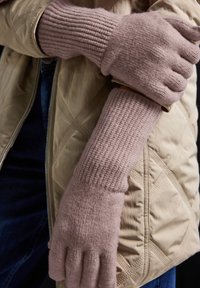 Rosa ribbade stickade handskar gjorda av mjukt material, åtsittande vid handleden, bärs av en person i en beige vadderad jacka.