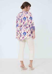 Lichtgewicht tuniek met bloemenpatroon in roomkleur, met blauwe en roze ontwerpen; gecombineerd met strakke beige broek en naakte bandjeshakken.