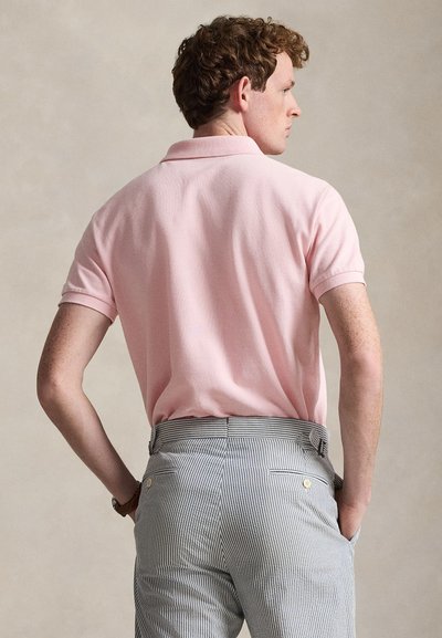 Άνδρας με την πλάτη γυρισμένη, φοράει πουκάμισο πόλο ανοιχτού ροζ με κοντά μανίκια και ψηλόμεσα παντελόνια με μπλε και λευκές ρίγες, με τα χέρια στις τσέπες.