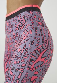Fioletowe legginsy z odważnym różowym i czarnym abstrakcyjnym wzorem, z czarnym paskiem w talii i kontrastującymi detalami ściegów.