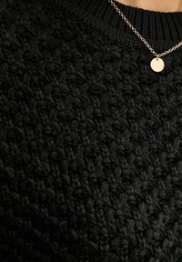 Zwarte gebreide trui met een gestructureerd, chunky patroon. Heeft een ronde halslijn en wordt gecombineerd met een delicate zilveren ketting met een kleine schijf.