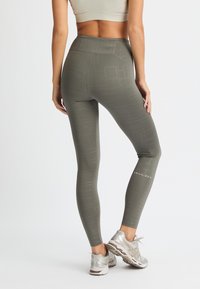 Leggings a vita alta di colore verde oliva con motivi geometrici e dettagli del logo sulla parte inferiore della gamba. Il tessuto sembra elastico e aderente.