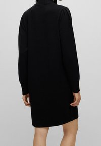 Robe noire en maille avec col montant, manches longues et poignets côtelés. Motif simple et droit, longueur mi-cuisse. Texture douce.