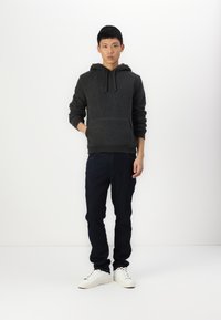 Sudadera de felpa gris oscuro con bolsillo frontal, capucha y cordones, combinada con jeans oscuros y zapatillas blancas sobre un fondo liso.