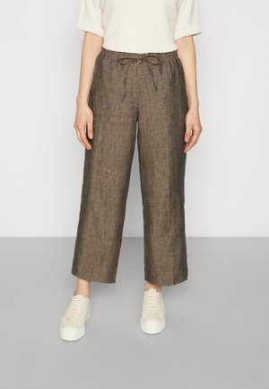 Personne portant un pantalon large en lin marron avec une taille à cordon et des baskets blanches, debout devant un fond uni.