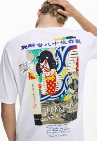 Valge T-särk, mille seljal on värvikas graafika, millel on Jaapani kabuki mask, lillemustrid ning sinised ja mustad tekstielemendid.