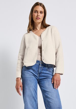 Cream-colored, fluffy-textured cropped jacket with a round neckline, snap buttons, and long sleeves. Paired with light blue jeans.
