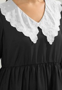 Musta mekko, jossa on valkoinen broderi-ristikolla kukkakuviot, kerätty vyötärö ja pehmeä, teksturoitu kangas.