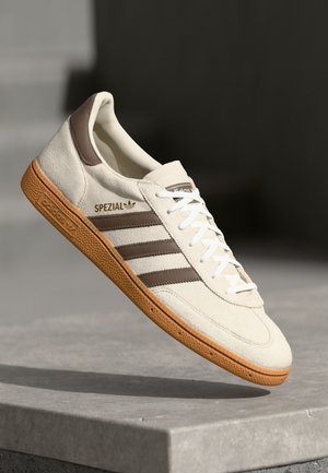 Zapatilla de ante crema con acentos de cuero marrón, tres franjas horizontales, cordones blancos y una suela de goma texturizada. Detalle de marca "SPEZIAL" en dorado.