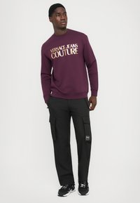 Felpa bordeaux con logo dorato "VERSACE JEANS COUTURE", abbinata a pantaloni cargo neri con più tasche e un accento testurizzato.