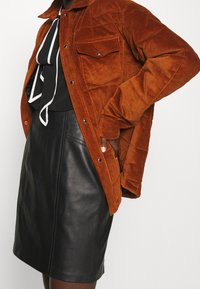 Samtjacke in verbranntem Orange mit zwei Brusttaschen, kombiniert mit einem schwarzen Lederrock. Weiche Textur, Druckknöpfe und eine lockere Passform.