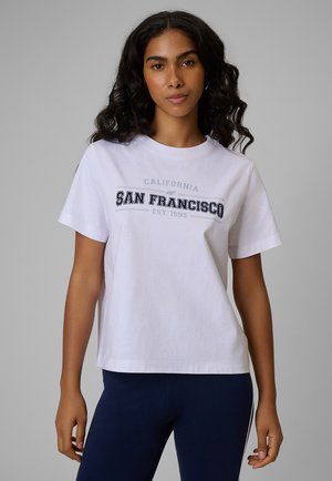 Kobieta o kręconych ciemnych włosach, ubrana w białą koszulkę z napisem „San Francisco California Est. 1995” oraz ciemnoniebieskie spodnie, stojąca na szarym tle.