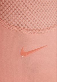 Nike urheiluvaatteet pehmeässä vaaleanpunaisessa kankaassa, jossa on teksturoitu verkkopaneeli ylhäällä. Esittää kiiltävää Nike-logoa selvästi näkyvillä.