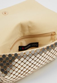 Bolso de mano metálico dorado con un patrón de espejos hexagonales, forrado con tela beige y que cuenta con un pequeño bolsillo con cremallera en el interior.