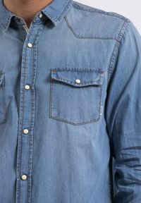 Chemise en denim bleu avec un devant boutonné, deux poches poitrine et des détails de couture visibles. Présente un col classique et une coupe décontractée.