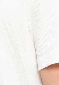 Κοντινό πλάνο του ώμου και του χεριού ενός ατόμου που φοράει ένα λευκό κοντομάνικο πουκάμισο με υφή βάφλας.