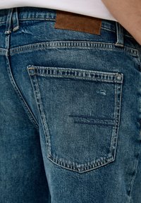 Dżinsy w średnim niebieskim kolorze z lekko wyblakłym wykończeniem. Zawierają skórzaną łatę w kolorze brązowym na pasku oraz tylną kieszeń ze szczegółowym przeszyciem.